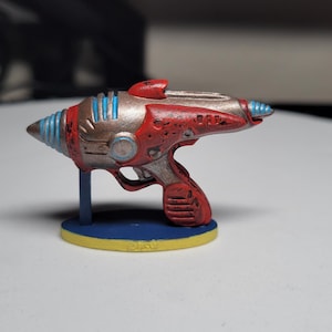 May include: A miniature ray gun figurine, primarily red with silver and blue accents. The toy gun is mounted on a blue stand with a yellow base. The design evokes a retro, science fiction aesthetic.