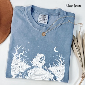 Puede incluir: Una camiseta color azul vaquero con un estampado gráfico blanco que muestra un esqueleto con vestido remando en un bote a través de un bosque con calabazas, estrellas y una luna creciente. La prenda tiene una etiqueta Comfort Colors y un collar dorado.