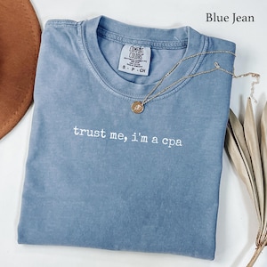 Puede incluir: Una camiseta color azul jean con cuello redondo y el texto bordado en blanco "trust me, i'm a cpa" en el pecho. La camiseta presenta una etiqueta de la marca Comfort Colors en el interior del cuello y se combina con un collar con colgante dorado.
