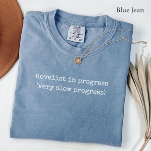May include: A blue jean colored t-shirt with a crew neckline and a Comfort Colors brand label at the inner collar. The shirt features white text that reads novelist in progress (very slow progress) and includes a gold chain necklace.
