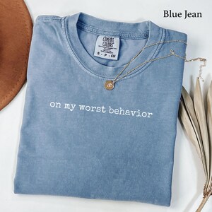 May include: A blue jean colored t-shirt with a crew neckline and the text "on my worst behavior" printed in white across the chest. The shirt features a Comfort Colors brand label at the inner neck and is paired with a gold chain necklace.