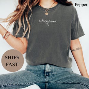 May include: A dark gray t-shirt with the word "entrepreneur" in white script, along with a small bow graphic. The shirt is paired with blue jeans. The word "Pepper" is printed in the top right corner.