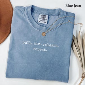 May include: A blue jean colored short-sleeve t-shirt with a crew neckline. The shirt features the white text "pull. aim. release. repeat." printed on the chest. A gold-toned necklace with a small circular pendant rests on the fabric.