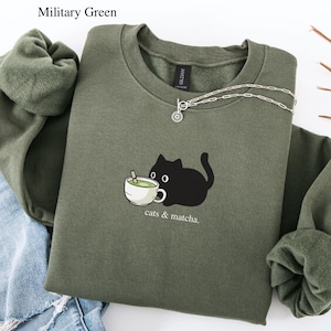 Könnte beinhalten: Militärgrünes Sweatshirt mit einer schwarzen Katzen-Grafik, die eine Matcha-Tasse hält. Der Text "cats & matcha" steht darunter. Eine silberne Halskette liegt auf dem Sweatshirt. Das Sweatshirt liegt neben einer Jeans.