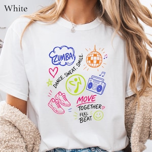 May include: A white t-shirt featuring a colorful graphic print with the text "ZUMBA", "DANCE. SWEAT. SMILE.", "MOVE TOGETHER", and "FEEL THE BEAT". The design includes a disco ball, a boombox, sneakers, a heart, a star, and a smiley face icon.