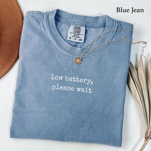 May include: A blue jean colored t-shirt with a crew neckline and short sleeves. The shirt features the white embroidered text low battery, please wait centered on the chest. A gold-toned necklace with a small circular pendant rests on the fabric.