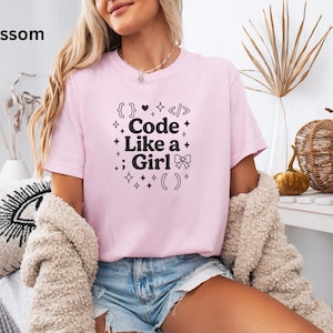 May include: Pale pink t-shirt with the slogan "Code Like a Girl" in black, incorporating code symbols and a bow. The shirt is short-sleeved with a round neck. The word "Blossom" is in the top left corner.