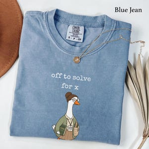 Op de afbeelding: Een blauw spijkerkleurig t-shirt met de witte tekst "off to solve for x" boven een afbeelding van een gans met een bril, een vest en een rok, die een boek en een etui vasthoudt. Een gouden ketting ligt op de kraag van het t-shirt.