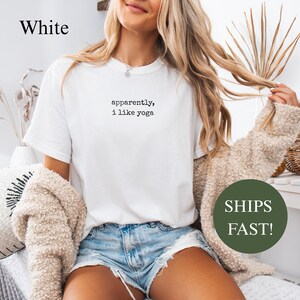 White t-shirt with "apparently, i like yoga" text, model wearing shorts and cardigan.
