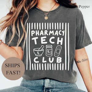 May include: A charcoal gray short-sleeve t-shirt featuring a white graphic print that reads PHARMACY TECH CLUB. The design includes icons of a mortar and pestle, a medicine bottle, and a bandage, all framed by vertical white lines.
