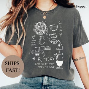 Może przedstawiać: Ciemnoszary t-shirt z krótkim rękawem z białymi rysunkami liniowymi przedstawiającymi narzędzia garncarskie, miskę, wazon i koło garncarskie. Tekst głosi: SHAPE YOUR STORY, FEEL THE PROCESS, CLAY, POTTERY, CRAFTED BY HAND, MADE TO HOLD.