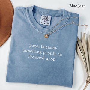 Op de afbeelding: Een blauw spijkerkleurig t-shirt met een ronde hals en een Comfort Colors-label in de binnenkraag. De voorkant is voorzien van witte tekst die luidt: yoga: because punching people is frowned upon. Een gouden ketting ligt op de stof.