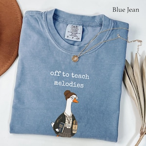 Puede incluir: Una camiseta color azul jean con el texto blanco off to teach melodies y un gráfico de un ganso con gafas y cárdigan, sosteniendo un libro de teoría musical y un portalápices. Un collar de cadena dorada descansa sobre el cuello.