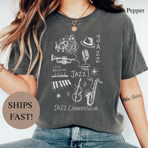 Op de afbeelding: Een antracietgrijs t-shirt met een witte lijntekening van jazz-gerelateerde elementen, waaronder een trompet, saxofoon, pianotoetsen, microfoon, hoed en oog. De tekst luidt "LIVE BREATHE JAZZ", "JAZZ" en "JAZZ CONNOISSEUR" in een handgetekende stijl.