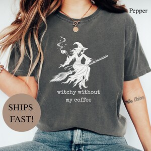 May include: A charcoal grey t-shirt featuring a white graphic of a witch flying on a broomstick while holding a steaming coffee cup. Below the graphic, the text reads, "witchy without my coffee." The shirt has short sleeves and a crew neckline.