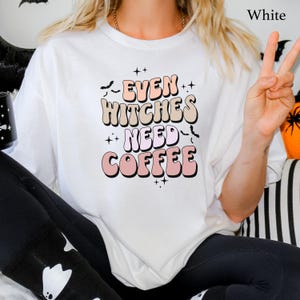 Peut inclure: Un t-shirt blanc à manches courtes avec le texte "EVEN WITCHES NEED COFFEE" écrit dans une police ondulée multicolore. Le motif comprend de petites silhouettes de chauves-souris noires et des étoiles à quatre branches autour du texte.