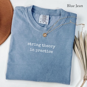 May include: A blue jean colored t-shirt with a crew neckline and the white embroidered text string theory in practice. The shirt features a Comfort Colors brand label at the inner neck and is displayed with a gold tone necklace pendant.
