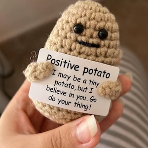 Puede incluir: Una figura de patata tejida a ganchillo en hilo beige con ojos de botón negros y una pequeña sonrisa. Sostiene un letrero blanco que dice "Positive potato" con un mensaje inspirador. Un artículo lindo, hecho a mano.