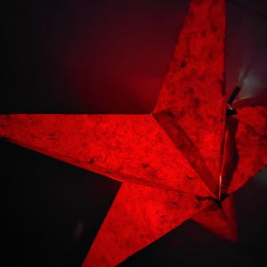 Może przedstawiać: Duża, czerwona dekoracja w kształcie pięcioramiennej gwiazdy. Gwiazda wykonana jest z teksturowanego materiału, prawdopodobnie papieru, i jest podświetlona, tworząc ciepłą poświatę. Ciemne tło podkreśla kształt i kolor gwiazdy.