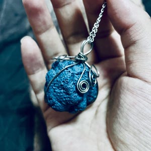 Może przedstawiać: Ręcznie robiony naszyjnik z teksturowaną, ciemnoniebieską zawieszką z kamienia. Kamień jest owinięty srebrnym drutem z ozdobnymi zawijasami. Zawieszka wisi na delikatnym srebrnym łańcuszku, tworząc unikatową i artystyczną biżuterię.