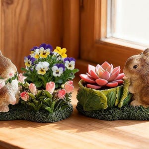 May include: Two brown bunny figurines, one holding a pink rose and the other next to a succulent. Between them are a pot of pansies and a succulent in a cabbage-shaped pot, all on a grassy base. Decorative garden decor.