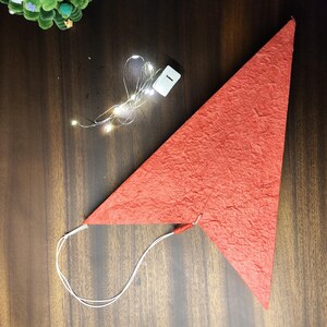 Może przedstawiać: Czerwona, tr&oacute;jkątna ozdoba w kształcie papierowej gwiazdy o teksturowanej powierzchni. Gwiazda jest przymocowana do białego sznurka i znajduje się obok sznura małych, ciepłobiałych diod LED i pojemnika na baterie. Ozdoba leży na ciemnej drewnianej powierzchni.