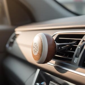 Könnte beinhalten: Ein Auto-Lufterfrischer mit einem kugelförmigen Holzdesign. Die Kugel hat eine helle Holzmaserung mit einem weißen Band und einem dekorativen, geschnitzten Zentrum. Er ist an einer Lüftungsöffnung im Auto befestigt.