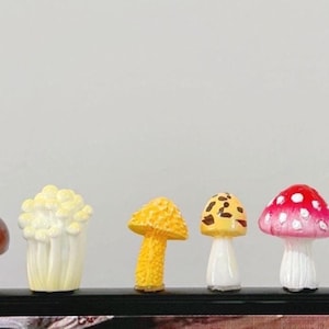 Op de afbeelding: Een verzameling van zeven miniatuur paddenstoelbeeldjes. De paddenstoelen variëren in kleur, waaronder bruin, geel, rood en wit. Sommige hebben vlekken of een textuur op de hoed. Ze zijn in een rij gerangschikt en tonen hun diverse ontwerpen.