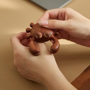 Könnte beinhalten: Handgefertigte Holz-Krabbenfigur mit detailliertem Design. Die Krabbe ist braun mit abgerundeten Merkmalen, einschließlich Augen und Scheren. Die Figur wird zwischen zwei Händen gehalten, was die Handwerkskunst und Größe der Krabbe zeigt.