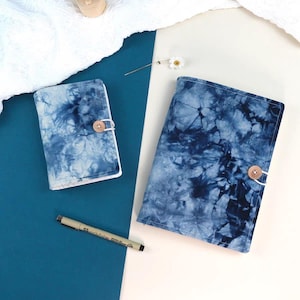 May include: Two blue tie-dye fabric journal covers with wooden button fastenings. One is smaller, and the other is larger. A black pen is on a blue surface, and a white cloth is in the background.