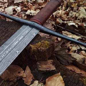 May include: A sword with a double-edged blade featuring a distinct wavy, layered metal pattern. The crossguard is made of dark, matte metal, and the handle is wrapped in brown leather, ending in a rounded pommel at the base of the grip.