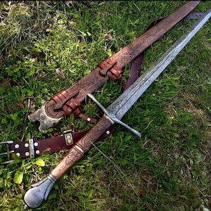 May include: A detailed image of a medieval sword with a decorative wooden handle and a silver blade. The sword rests on green grass next to a brown leather sheath and belt, both with intricate carvings and metal accents.