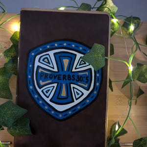 Puede incluir: Un libro marrón con un diseño de escudo azul y blanco y el texto "PROVERBIOS 30:5". El libro está decorado con hiedra artificial y luces de hadas.