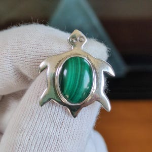 May include: A silver turtle-shaped pendant featuring a central oval-cut malachite gemstone with distinct dark green banding. The metal frame is polished and smooth, forming the head, four flippers, and a tail, with two small circular indentations on the head.