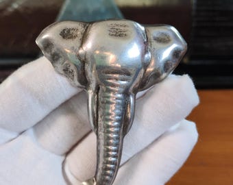 May include: A silver-toned metal brooch shaped like an elephant head. The piece features a polished finish with detailed textures on the trunk and ears, showcasing a symmetrical design with prominent tusks and a curved trunk pointing downward.