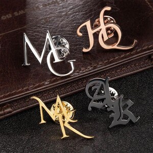 Op de afbeelding: Vier metalen initialen-spelden in zilver, roségoud, goud en zwart. De spelden tonen de letters MG, HQ, AK en NA in verschillende gestileerde lettertypen. De spelden worden gepresenteerd op een bruin lederen oppervlak en een zwart lederen oppervlak.