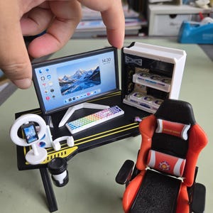 Puede incluir: Un set de escritorio de juego en miniatura que incluye un escritorio negro con detalles amarillos, un monitor de computadora con un paisaje de montaña, una torre de PC blanca, un teclado arcoíris, una luz de anillo con teléfono, auriculares blancos y una silla roja y negra.