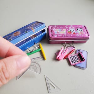 Könnte beinhalten: Miniatur-Schulmaterialien, darunter zwei Federmäppchen, eines in Blau und eines in Rosa, mit Cartoon-Designs. Außerdem kleine Lineale, ein Winkelmesser, ein Taschenrechner, ein Stift und ein Tacker. Auf dem rosa Federmäppchen steht "Dream Animal".