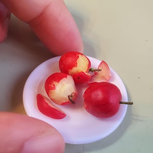 May include: Miniature red apples, some partially peeled, rest on a small white plate. The apples have a realistic appearance with a red exterior and a light-colored interior. A finger is seen near the plate.