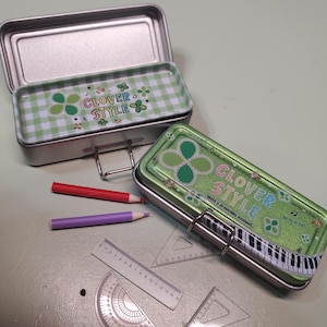 May include: Two metal pencil cases with the text "CLOVER STYLE" and a green clover design. The cases are open, revealing a green and white checkered pattern. Also, two pencils and a ruler are visible.