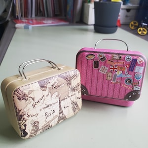Puede incluir: Dos pequeñas maletas con temática de viaje. Una es beige con un diseño de mapa del mundo y etiquetas de destinos de viaje. La otra es rosa, decorada con pegatinas de viaje, incluyendo "Venezia". Ambas tienen asas plateadas.