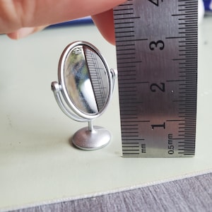 May include: A miniature silver-coloured oval tabletop mirror with a swivel frame and a round base. A metal ruler is positioned next to the mirror, showing the item stands approximately 3.8 cm tall from the base to the top of the frame.