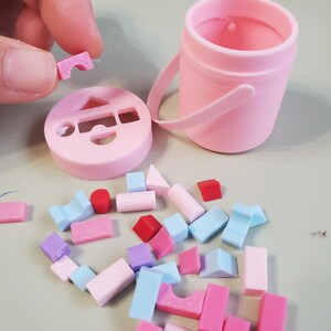 May include: A light pink plastic bucket with a matching lid featuring various geometric cutouts, including a triangle, rectangle, and semicircle. A collection of small, multicolored geometric shapes in red, pink, purple, and blue are scattered nearby.