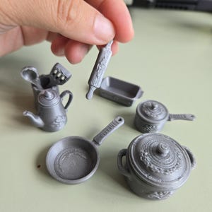 Peut inclure: Un ensemble d'ustensiles de cuisine miniatures gris comprenant un rouleau à pâtisserie, un moule à pain, une casserole avec couvercle, une poêle, une théière et un pot à ustensiles avec cuillère et spatule, tous avec une finition imprimée en 3D.
