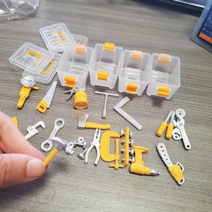 Op de afbeelding: Een verzameling miniatuur speelgoedgereedschap van plastic, waaronder een hamer, zaag, bijl, tang, moersleutel en boormachine, vergezeld van vier kleine doorzichtige opbergbakjes met oranje sluitingen en deksels in geel en zilver.