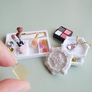 May include: A collection of miniature vanity accessories including a white tray with makeup brushes, small perfume bottles, and a gold chain. Nearby is a black eyeshadow palette, a white ornate decorative piece, and a pendant with a pink gem.