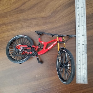 Könnte beinhalten: Ein rotes und schwarzes Mountainbike-Modell mit schwarzen Rädern und einer gelben Vorderradaufhängung. Das Fahrrad befindet sich neben einem Lineal und misst etwa 25 cm in der Länge. Der Text "MODELBICICLE" ist auf dem Rahmen zu sehen.