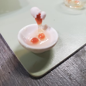 May include: A miniature ceramic bowl with a cracked eggshell and dripping yolk. Two orange yolks sit in the white bowl. The egg is positioned above the bowl, with the yolk dripping down. The background is a light green surface.