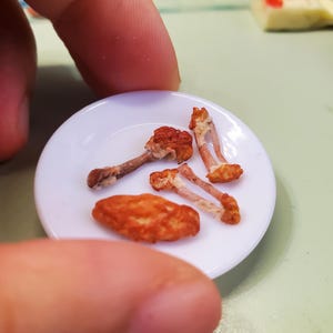 May include: A small white plate holding miniature food items, including what appears to be fried chicken and mushrooms. The food items are brown and orange. The plate is held by a person's fingers.
