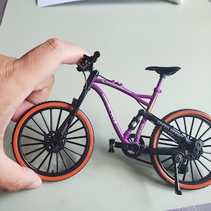 Könnte beinhalten: Ein Miniatur-Fahrradmodell in Lila mit schwarzen Rädern und leuchtend orangefarbenen Reifen. Der Rahmen trägt den Schriftzug Speed Sprint und umfasst einen schwarzen Sattel, einen Lenker und einen Ständer. Das Design enthält eine Hinterradfederung.